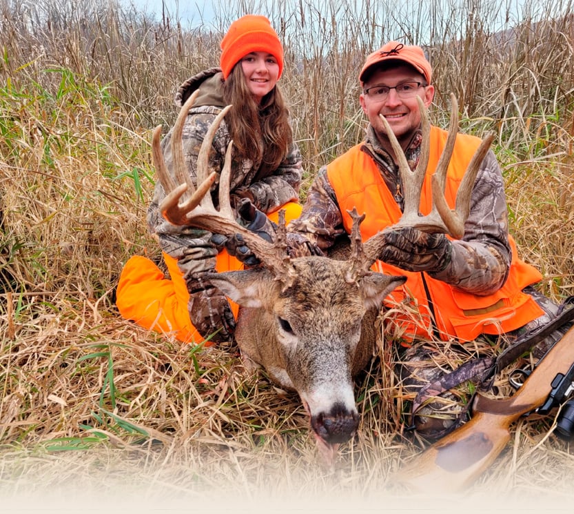 A father and daughter sit in tall, dry grass next to a harvested whitetail buck.